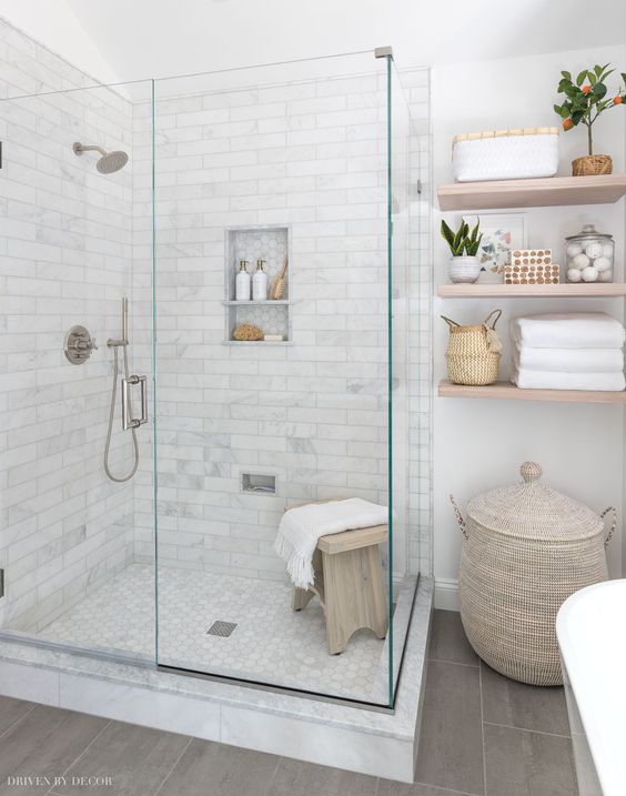 a neutral farmhouse bathroom with floating shelves, a basket with a lid, a shower space enclosed in glass and a wooden stool