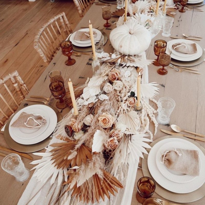 a gorgeous boho Thanksgiving tablescape with gold placemats and white porcelain, a lush table runner made of pampas grass, fronds, a white pumpkins, pink and white blooms