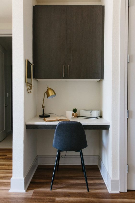a small and chic home office nook with a built in cabinet, a built in desk, a navy chair, a brass lamp and a mirror