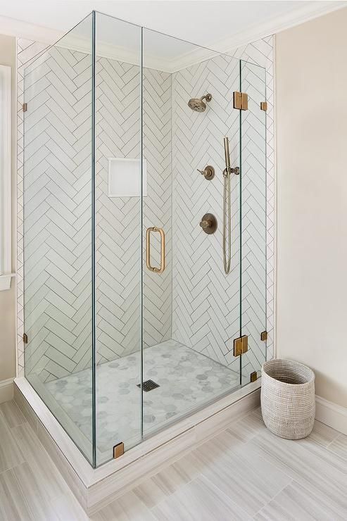 a shower space clad with herringbone and marble tiles, brass fixtures and a glass door with a brass handle