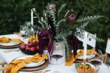 14 a bright modern Thanksgiving table with greenery and deep purple blooms, yellow napkins, woven chargers and gold cutlery rocks