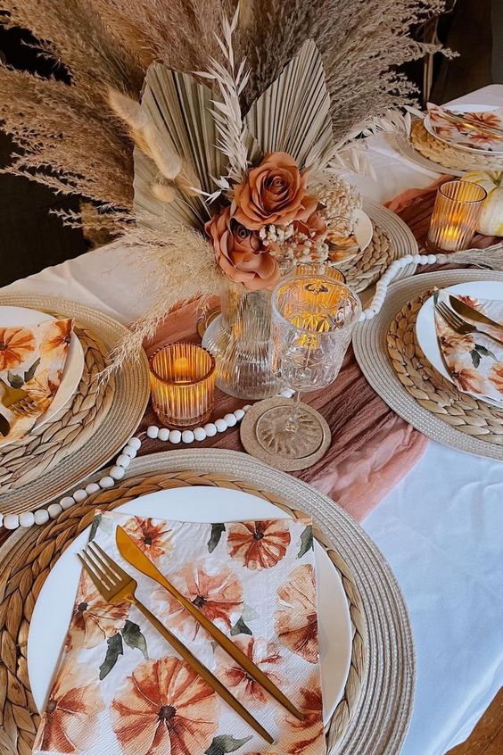 a pretty boho Thanksgiving table setting with a rust runner, a large rust rose and dried frond centerpiece, wooden beads, woven placemats