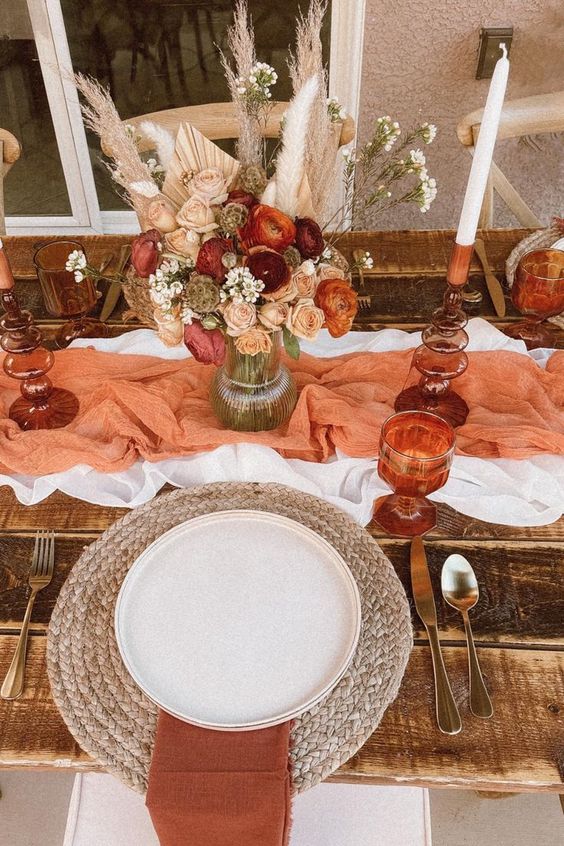a warm-colored boho Thanksgiving tablescape with a white and orange table runner, a dried flower and pampas grass bouquet, a woven placemat and rust napkins