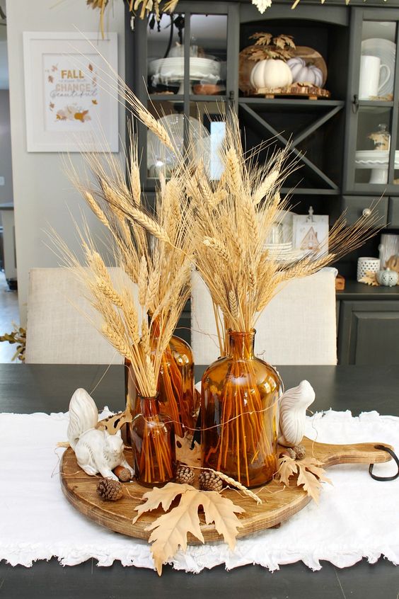 a boho Thanksgiving centerpiece of a cutting board, leaves and pinecones and apothecary bottles with wheat is a lovely idea