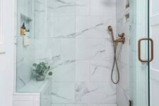 20 a refined shower space with white marble and penny tiles, glass doors and brass fixtures plus greenery in a vase