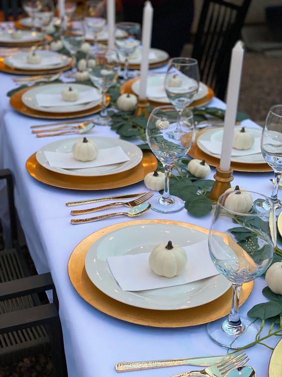 a modern Thanksgiving tablescape with gold chargers and candleholders plus cutlery, white porcelain and pumpkins, leaves and candles