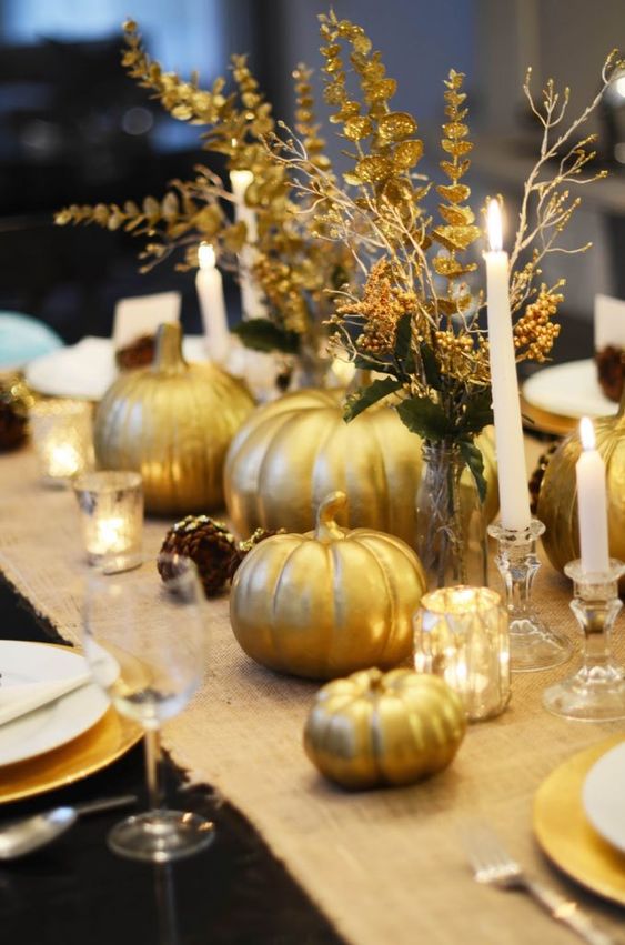 a pretty gold Thanksgiving tablescape with a burlap runner, gold pumpkins, gold candleholders, pinecones and candles, gilded leaves