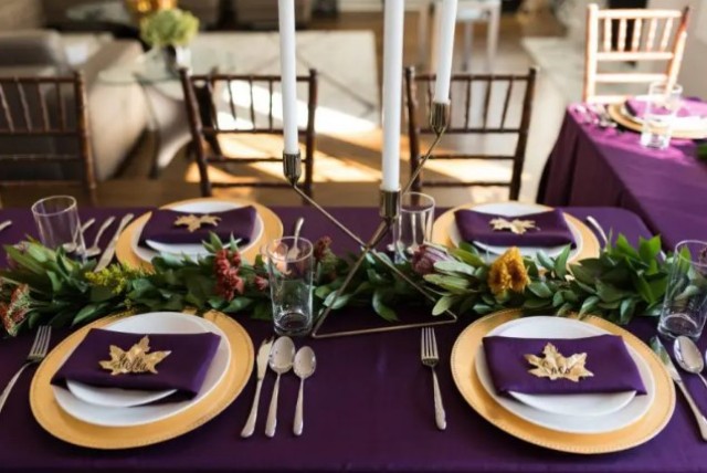 a gorgeous purple Thanksgiving tablescape with a purple tablecloth and napkins, a greenery and bright bloom runner, tall candles, gold touches and fall leaves