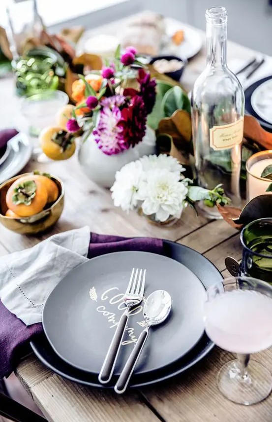 a laid back and rustic Thanksgiving tablescape with greenery, bold purple blooms, fruits, black plates and purple napkins