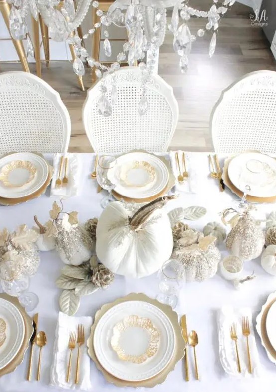 a refined white and gold Thanksgiving tablescape with white embellished pumpkins, white leaves, gold chargers and cutlery plus a crystal chandelier over the table