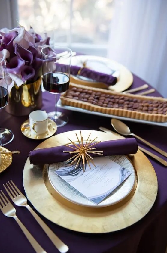 a modern glam Thanksgiving tablescape with a purple tablecloth, napkins and callas, gold charrgers and cutlery and a vase