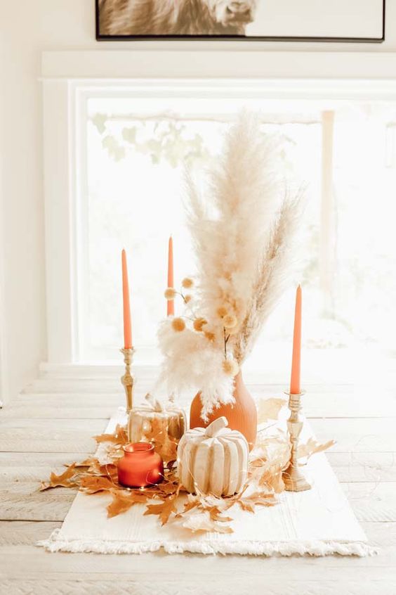 a neutral boho Thanksgiving centerpiece of wooden pumpkins, a terracotta vase and candles, dried grasses, lights and leaves