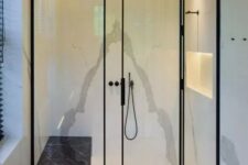 28 a shower space done with white stone tiles, a lit up niche, black frame glass doors and black fixtures plus a black marble bench