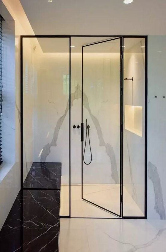 a shower space done with white stone tiles, a lit up niche, black frame glass doors and black fixtures plus a black marble bench