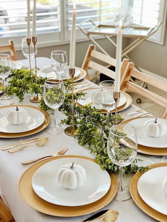 a stylish Thanksgiving table setting with gold chargers and cutlery, candleholders, mini white pumpkins and greenery