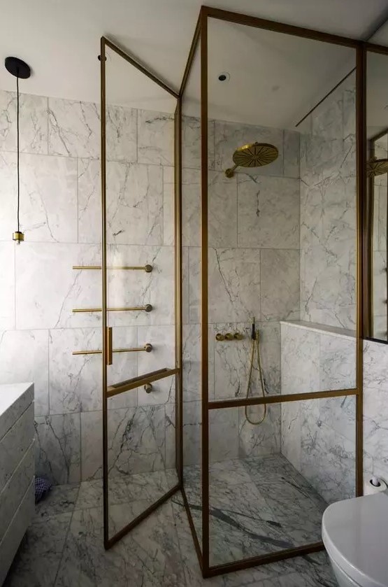 a chic and refined bathroom clad with white marble tiles, with a shower space and with brass frames doors and brass fixtures