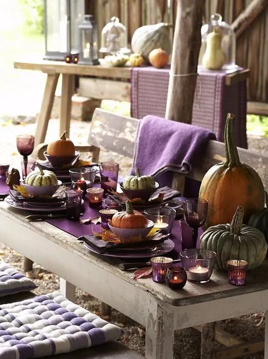 a rustic Thanksgiving tablescape with a purple table runner, napkins, glasses and candle holders, large natural pumpkins and towels