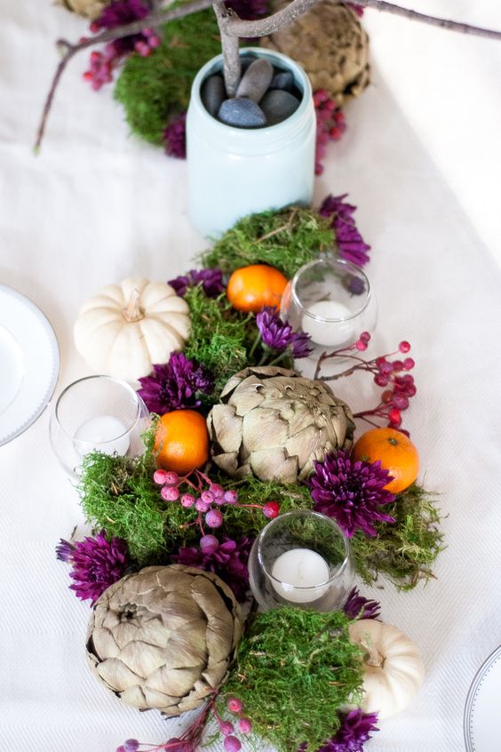 an eye-catchy Thanksgiving centerpiece with moss, white pumpkins, artichokes, berries, citrus and some candles plus purple blooms