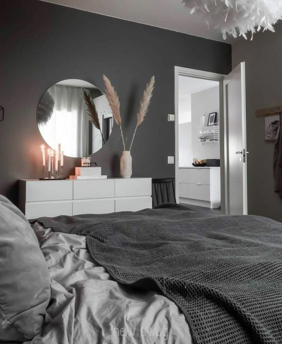 a Scandinavian bedroom with black walls, a bed with monochromatic bedding, a white dresser, a round mirror and candles