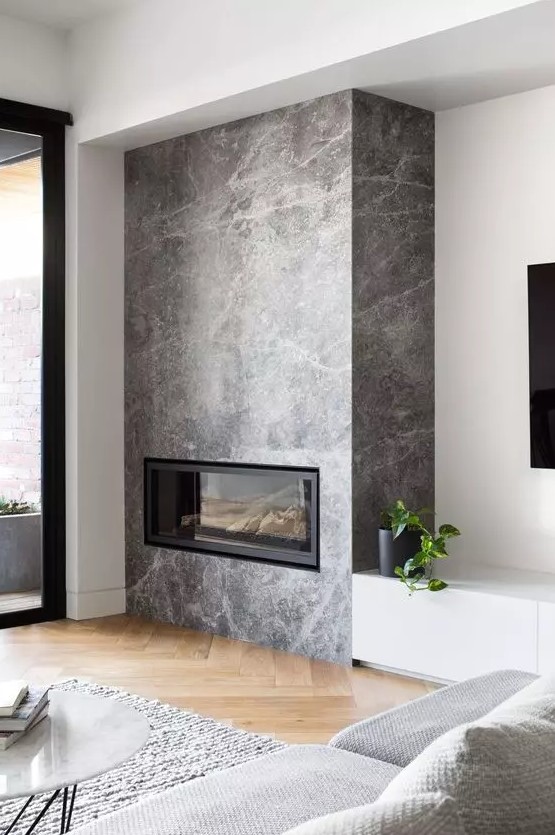 a Scandinavian living room with a built-in fireplace in grey stone, a grey sofa and a rug, a wihte TV unit and a marble coffee table