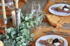 a beautiful Thanksgiving table setting with orange placemats and napkins, greenery table runners, candles and boards