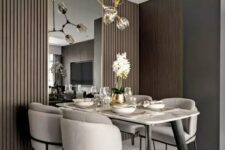 a beautiful contemporary dining space with a marble top table, neutral chairs, a large mirror, taupe wooden panels and a catchy chandelier
