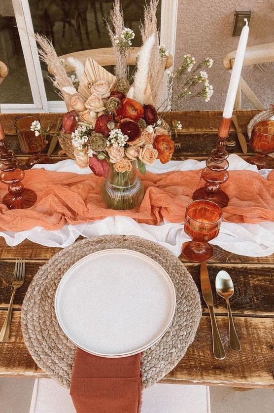 a beautiful rust Thanksgiving tablescape with a white and orange table runner, a dried flower and pampas grass bouquet, a woven placemat and rust napkins