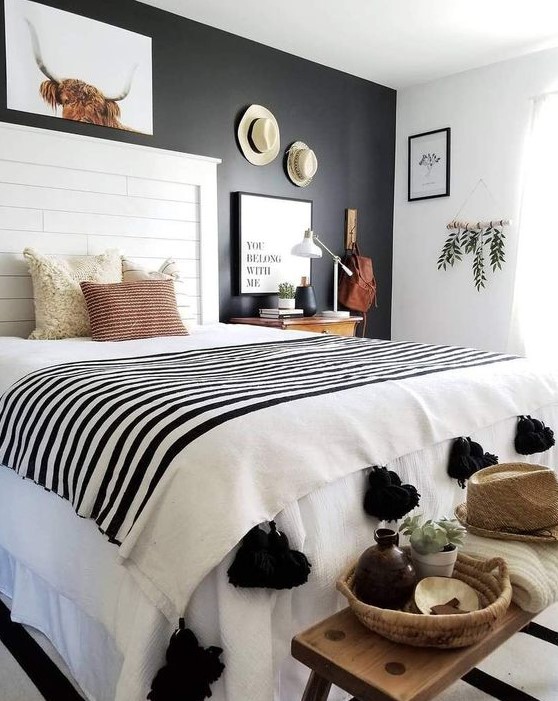 a black and white boho bedroom with a black statement wall, a white bed, a wooden bench, a gallery wall with hats and a tassel blanket