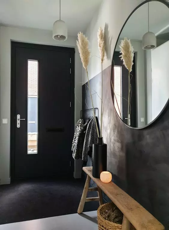 a black and white boho entryway with a black accent wall, door and a rug, a stained bench, pampas grass, a round mirror and a pendant lamp