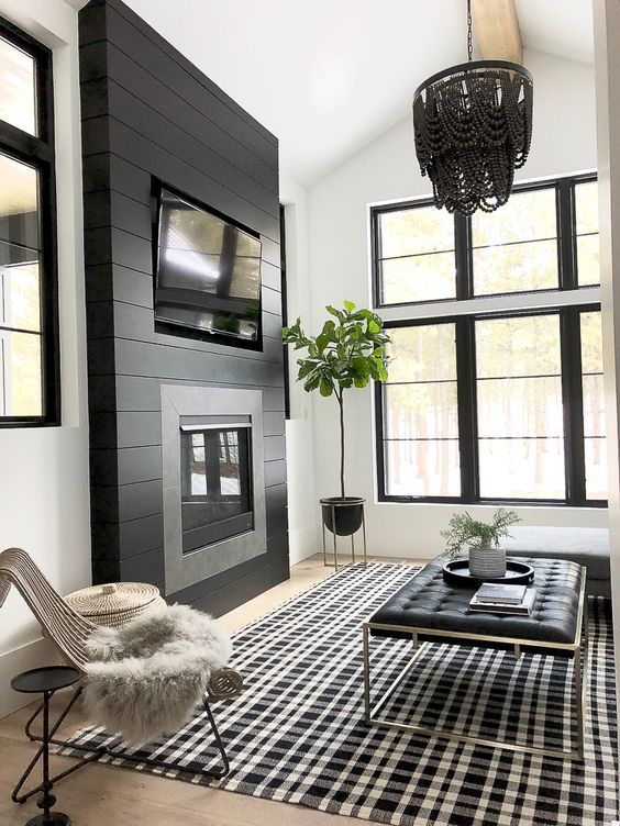 a black and white living room with a shiplap wall and a built-in fireplace, a plaid rug, a leather ottoman and a rattan chair