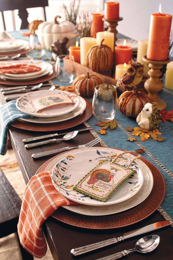 a bright Thanksgiving tablescape with orange and blue napkins and a table runner, pillar candles, vintage cards and fall leaves and pumpkins