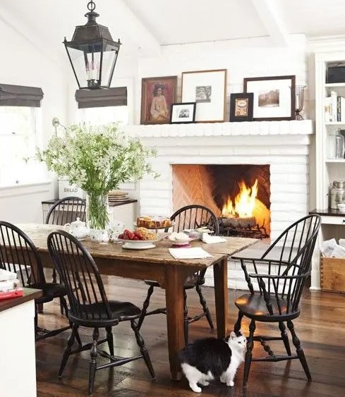 a bright white farmhouse dining space with a white brick fireplace, a wooden table and black chairs, artworks and lanterns