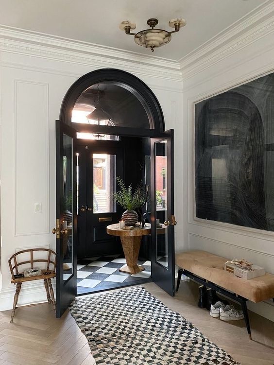 a chic black and white entryway with a neutral bench, a stained chair, a black and white rug and black doors