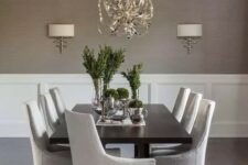 a chic modern dining room with taupe grasscloth wallpaper walls, creamy paneling, a black table and neutral chairs, a lovely chandelier