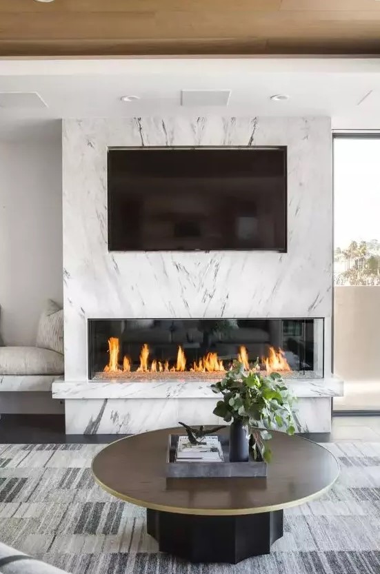 a chic modern space with a fireplace clad with white marble and a TV, a built in bench, a round table and a statement rug