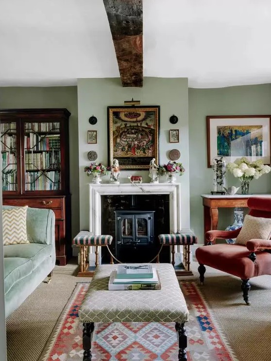 a cozy and welcoming vintage living room with light green walls, a hearth, a green sofa, a red chair, printed ottomans, a gallery wall