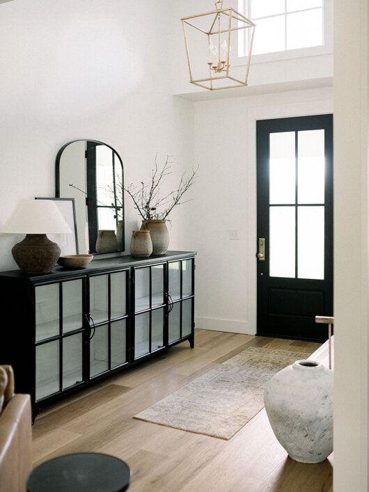 a farmhouse entryway with a black storage unit, a black door, a neutral rug, a large vase, an arched mirror and a gilded chandelier