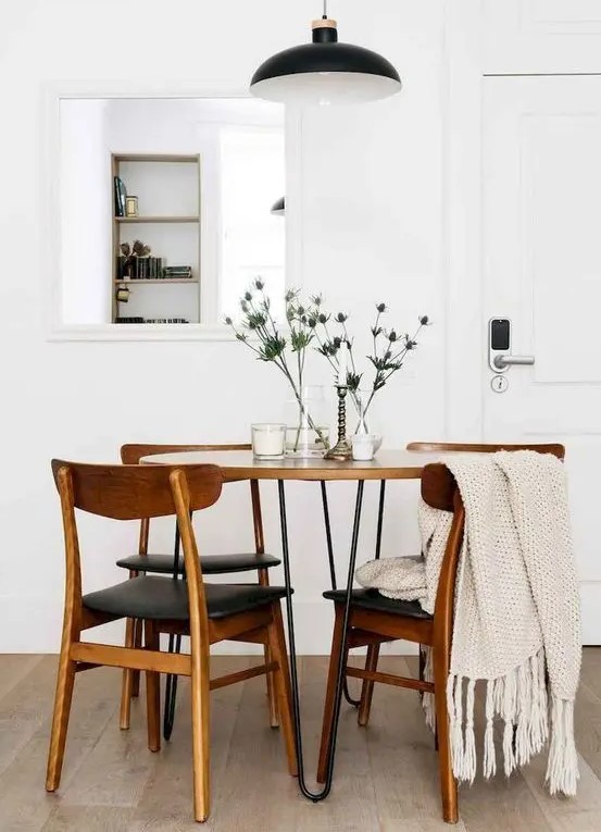 a laconic mid century modern dining room with a mirror, a round hairpin leg table, black chairs, a black pendant lamp and thistles