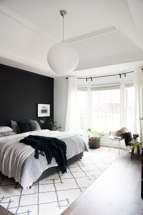 a light-filled bedroom with a black accent wall, a bed with black and white bedding, a bay window, a neutral chair and greenery