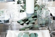 a minimal Thanksgiving tablescape with white square plates, a greenery runner and pillar candles on the table, refined glasses