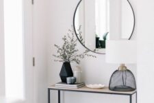 a minimal and clean black and white entryway with a sleek console table, a round mirror, a table lamp and a geo vase