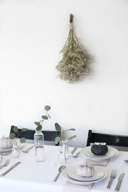 a minimalist Thanksgiving table setting with a white tablecloth and grey napkins, white porcelain and grey and black pumpkins plus greenery in vases