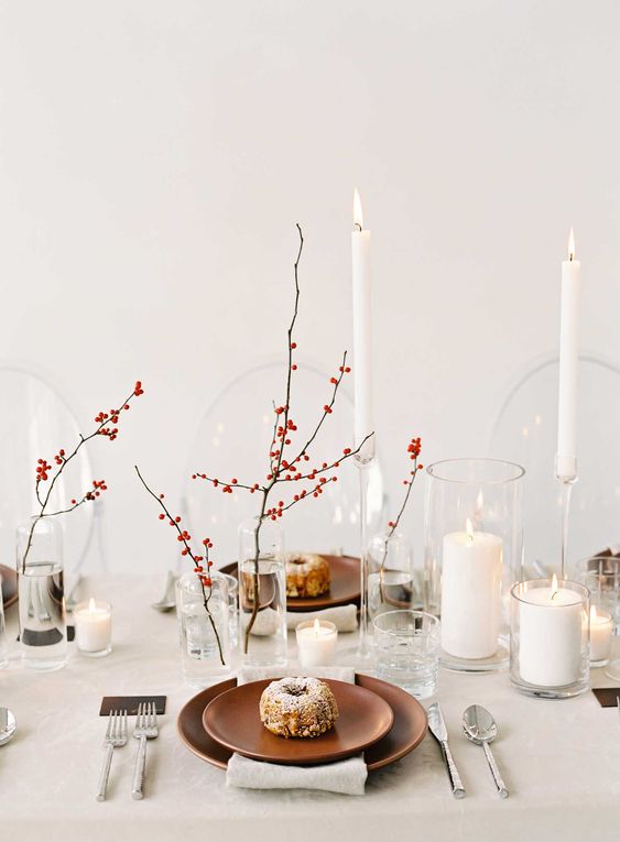 a minimalist Thanksgiving tablescape done in neutrals, with berries in vases, tall and thin and pillar candles, rust colored plates and silver cutlery