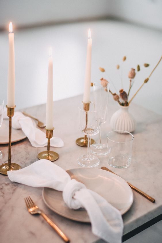 a minimalist Thanksgiving tablescape with neutral porcelain, gold cutlery and gold candleholders, a white vase with grasses