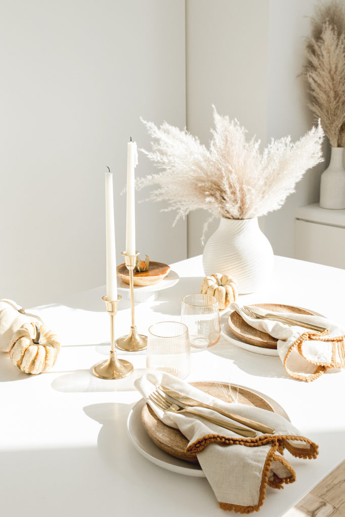 a minimalist meets boho Thanksgiving tablescape with a white vase with grasses, neutral porcelain and wooden plates, pumpkins and candles