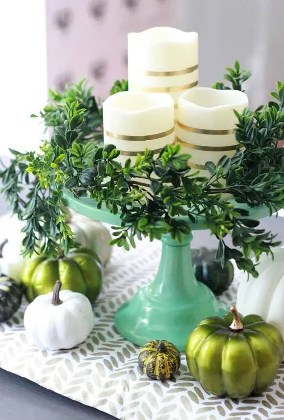 a modern Thanksgivving centerpiece of a green stand with greenery, striped candles, white and green pumpkins and a printed runner