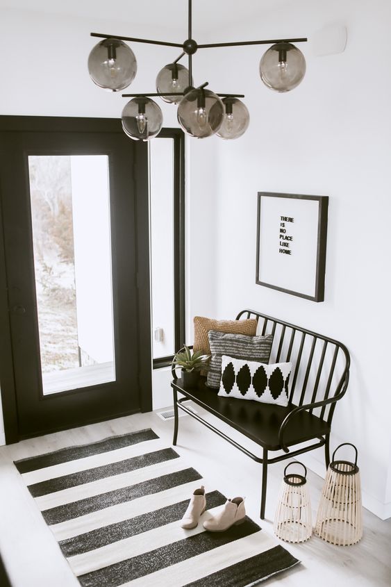 a modern black and white foyer with a black glass door, a black bench and a striepd rug, an artwork and lanterns