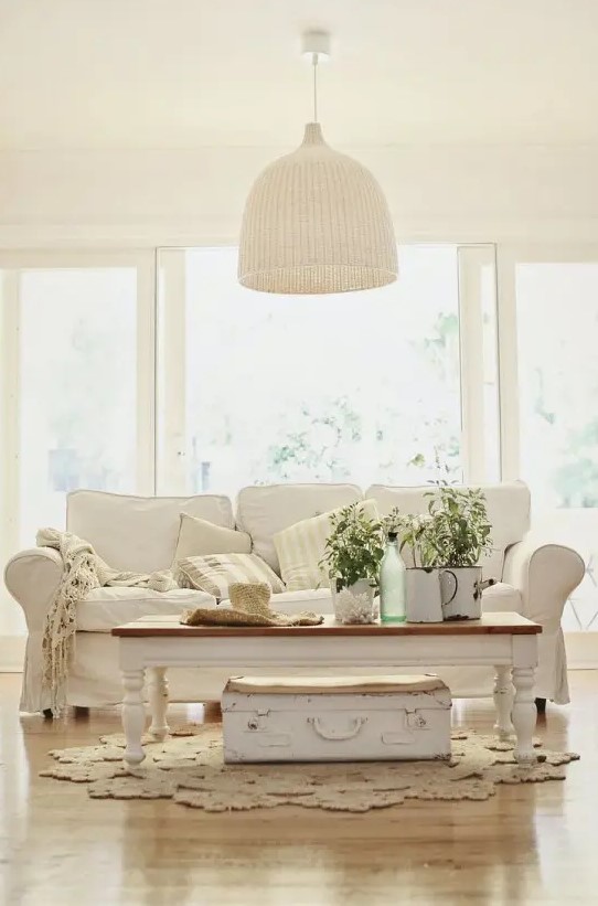 a neutral Provence styled living room with a white sofa, a low coffee table and a suitcase, a woven pendant lamp and some potted greenery