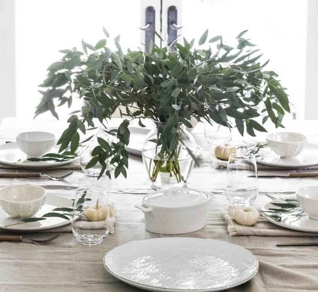 a neutral minimalist Thanskgiving tablescape with a lush greenery centerpiece in a clear vase, white porcelain, mini white pumpkins and neutral napkins