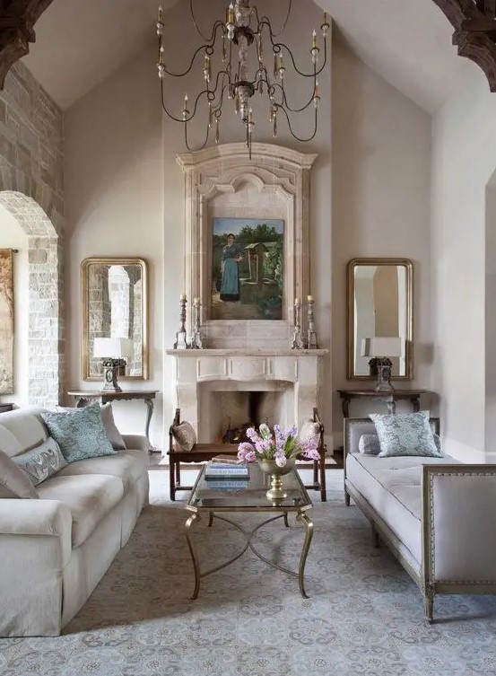 a neutral vintage living room with a fireplace, a refined chandelier, two mirrors, a couple of sofas and a glass table